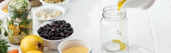 Cropped view of woman pouring oil in empty glass jar on wooden white table, panoramic shot — Stock Photo