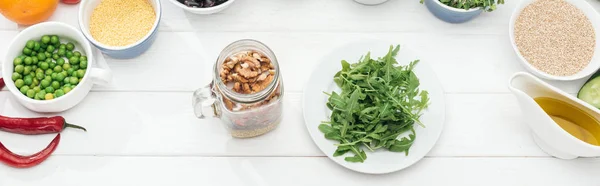 Top view of glass jar with nuts near plate with green arugula on wooden white table, panoramic shot — Stock Photo