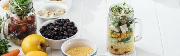 Ingredienti in ciotola vicino a vasi di vetro con insalata su tavolo bianco di legno, colpo panoramico — Foto stock