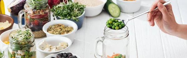 Cropped view of woman adding green peas in glass jar on wooden white table, panoramic shot — Stock Photo