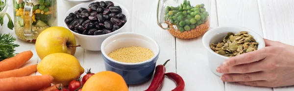 Cropped view of woman adding pumpkin seeds in glass jar on wooden white table, panoramic shot — Stock Photo