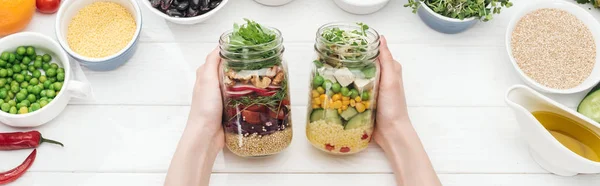 Cropped view of woman holding jars with tasty salad on wooden white table , panoramic shot — Stock Photo