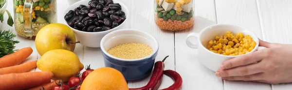 Cropped view of woman holding corn in bowl on wooden white table, panoramic shot — Stock Photo