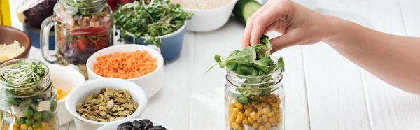 Cropped view of woman adding greens in glass jar on wooden white table, panoramic shot — Stock Photo