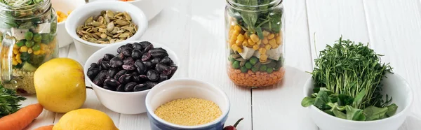 Greens in bowl near fruits and vegetables in glass jar on wooden white table, panoramic shot — Stock Photo