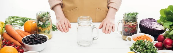 Vue recadrée d'une femme dans un tablier debout près d'un bocal en verre vide sur une table en bois isolée sur une photo panoramique blanche — Photo de stock