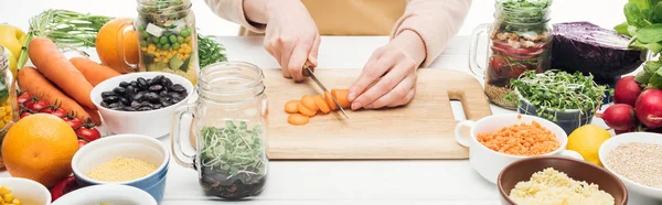 Cropped view of woman in apron cutting carrots on wooden table isolated on white, panoramic shot — Stock Photo