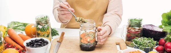 Cropped view of woman in apron adding pumpkin seeds to glass jar with beans on wooden table isolated on white, panoramic shot — Stock Photo