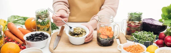 Cropped view of woman in apron adding pumpkin seeds from bowl in glass jar on wooden table isolated on white, panoramic shot — Stock Photo