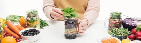Partial view of woman in apron holding glass jar with salad and green leaves on wooden table isolated on white, panoramic shot — Stock Photo