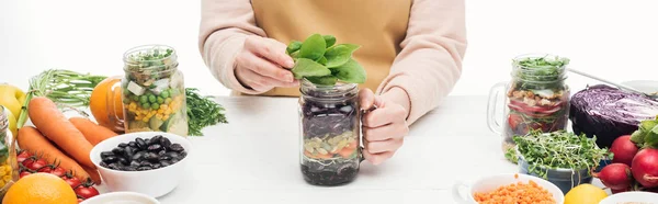 Teilansicht einer Frau in Schürze mit Glasgefäß mit Salat und grünen Blättern auf Holztisch isoliert auf weißem, panoramischen Bild — Stockfoto