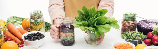 Cropped view of woman in apron holding glass jar with salad and green leaves on wooden table isolated on white, panoramic shot — Stock Photo