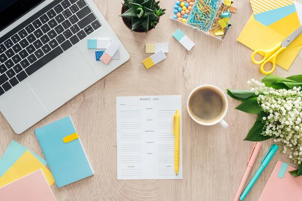 Vista superior da lista semanal, xícara de café, artigos de papelaria, laptop e flores na mesa de madeira — Fotografia de Stock