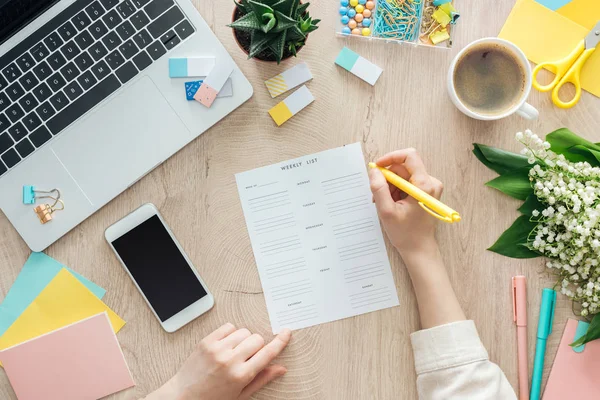 Vista recortada de la mujer que sostiene la pluma en la lista semanal, trabajando en la mesa de madera con taza de café, papelería, computadora portátil y flores - foto de stock