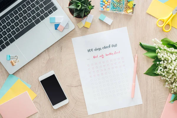 Vue du dessus de la liste de vérification de 100 jours avec smartphone, ordinateur portable et papeterie sur table en bois — Photo de stock