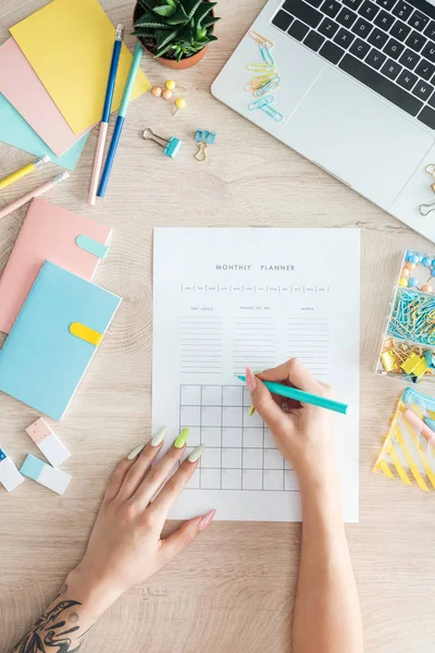 Cropped view of woman sitting behind wooden table with laptop and stationery, writing notes in monthly planner — Stock Photo