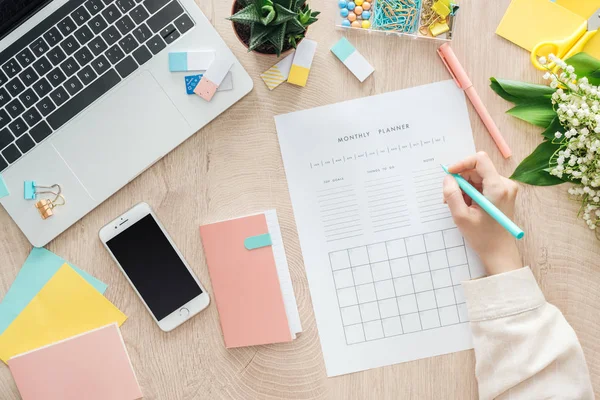 Cropped view of woman sitting behind wooden table with smartphone, laptop and stationery, writing notes in monthly planner — Stock Photo