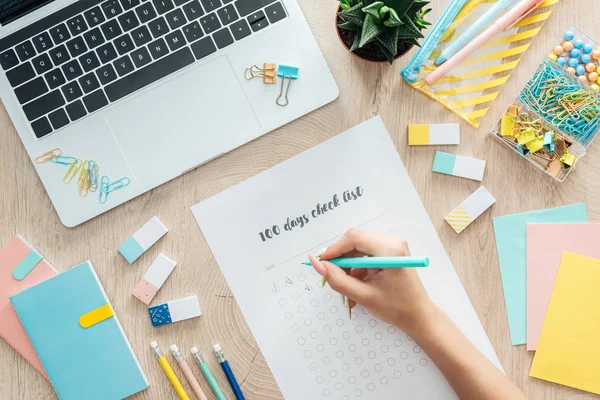 Vista ritagliata di donna scrittura note in 100 giorni lista di controllo, seduto dietro tavolo in legno con cancelleria, fiori e laptop — Foto stock