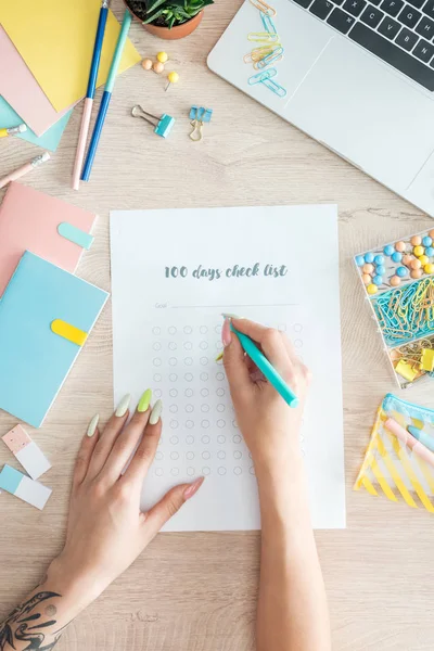 Cropped view of woman sitting behind wooden table with laptop and stationery, writing notes in 100 days check list — Stock Photo