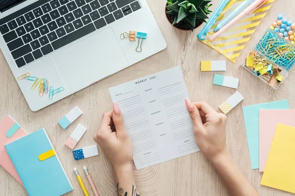 Cropped view of woman sitting behind wooden table with laptop and stationery, holding in hands monthly list — Stock Photo