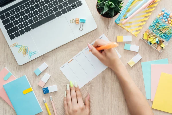 Vista recortada de la mujer escribiendo notas en planificador, sentado detrás de la mesa de madera con el ordenador portátil, papelería y planta - foto de stock