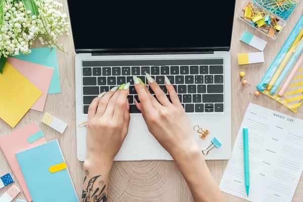 Cropped view of woman texting on laptop keyboard, working behind wooden table with flowers and stationery — Stock Photo