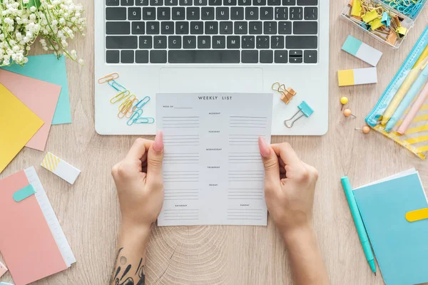 Cropped view of woman sitting behind wooden table with laptop, stationery and holding weekly list in hands — Stock Photo