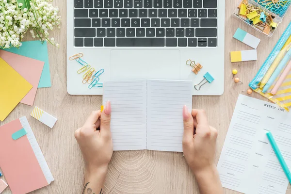 Cropped view of woman sitting behind wooden table with laptop, weekly list, stationery and holding notepad in hands — Stock Photo