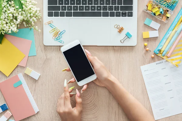 Cropped view of woman sitting behind wooden table with laptop, weekly list, stationery and holding smartphone in hands — Stock Photo