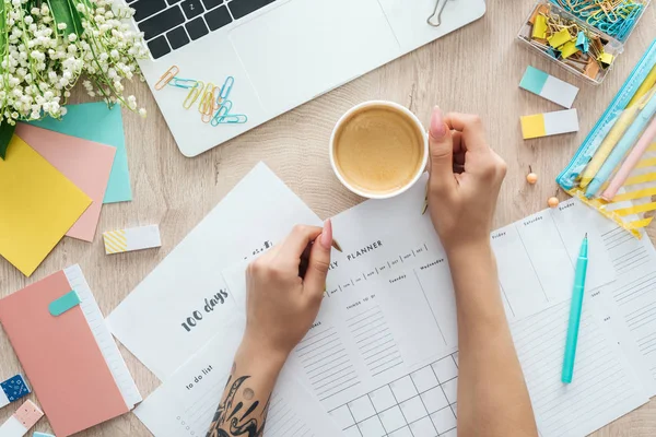 Cropped view of woman sitting with cup of coffee behind wooden table with planners, stationery and laptop — Stock Photo