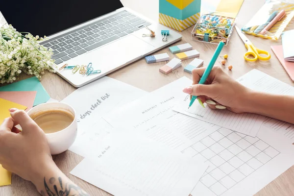 Vue recadrée de la femme tenant une tasse de café à la main, assise derrière une table en bois avec ordinateur portable et papeterie, écrivant dans des planificateurs de papier — Photo de stock