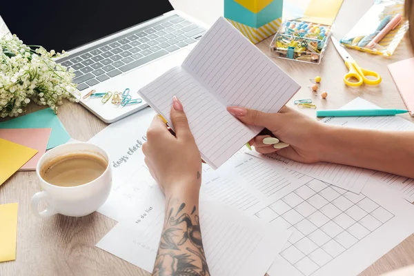 Vista ritagliata della donna che tiene il blocco note in mano, lavorando dietro il tavolo in legno con tazza di caffè, cancelleria, laptop e fiori — Foto stock