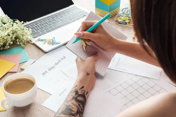 Cropped view of woman writing notes in notepad, sitting behind wooden table with laptop, planners and stationery — Stock Photo