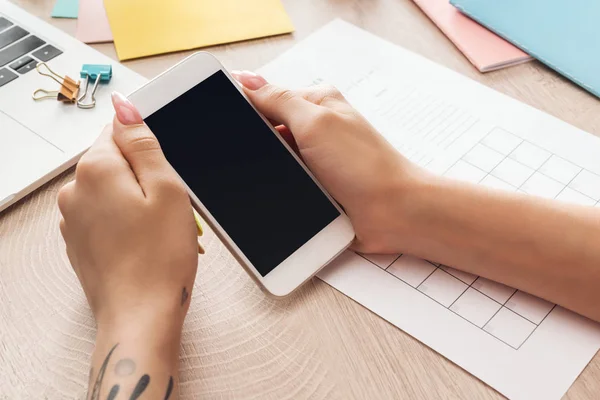 Vista recortada de la mujer sentada detrás de una mesa de madera con computadora portátil y papelería, sosteniendo el teléfono inteligente en las manos — Stock Photo