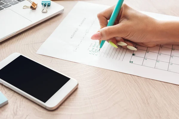 Cropped view of woman writing notes in planner, sitting behind wooden table with laptop and smartphone — Stock Photo