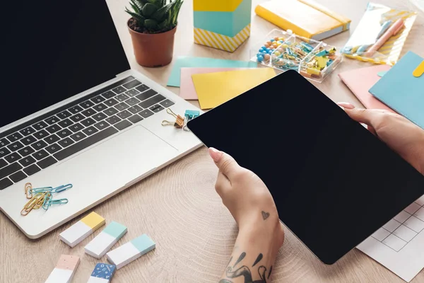 Ausgeschnittene Ansicht einer Frau, die ein digitales Tablet in der Hand hält und hinter einem Holztisch mit Laptop und Schreibwaren sitzt — Stockfoto