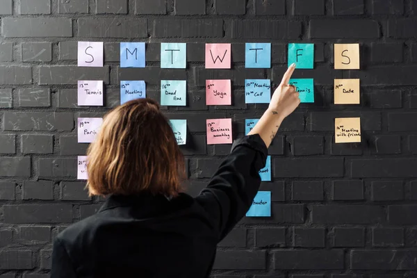 Vista posterior de la mujer apuntando con el dedo a las notas de etiqueta de colores en la pared de ladrillo negro - foto de stock