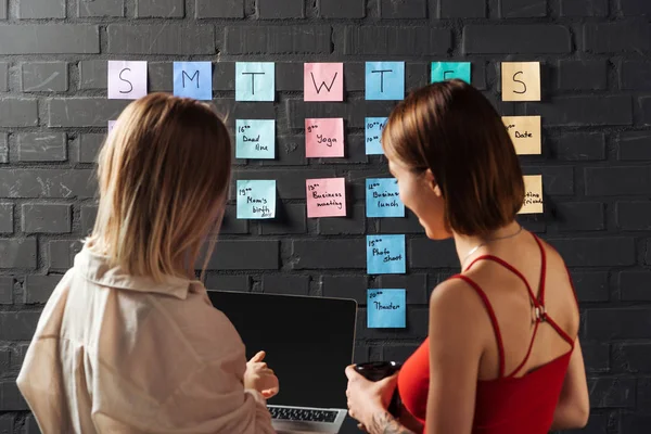 Back view of two women standing near colorful sticker notes on black brick wall, looking at laptop — Stock Photo