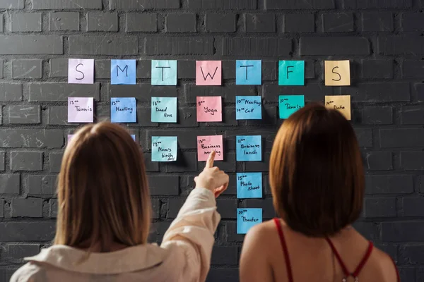 Vue arrière de deux femmes debout près de notes autocollantes colorées et pointant du doigt sur un mur de briques noires — Photo de stock