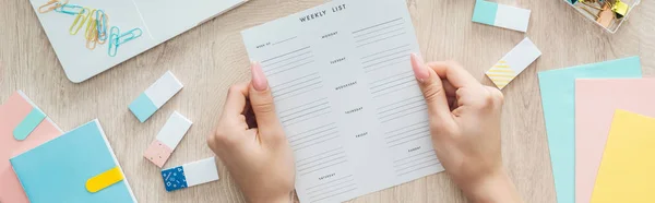 Cropped view of woman holding weekly list in hands over wooden table with stationery — Stock Photo