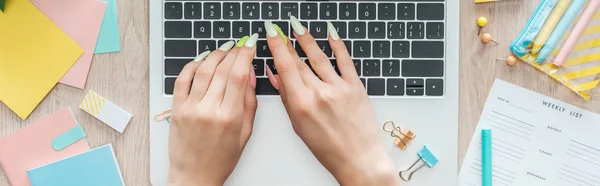 Обрізаний вид жінки, що друкує на клавіатурі ноутбука, сидить за дерев'яним столом з канцелярськими приладдям і щотижневим списком — стокове фото