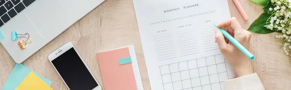 Cropped view of woman writing notes at monthly planner, sitting behind wooden table with stationery, notepad and smartphone — Stock Photo