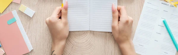 Vista recortada de la mujer sosteniendo bloc de notas en las manos sobre la mesa de madera con lista semanal y papelería - foto de stock