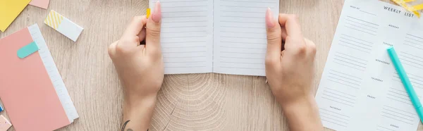 Vue recadrée de la femme tenant un bloc-notes dans les mains sur une table en bois avec liste hebdomadaire et papeterie — Photo de stock