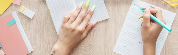 Cropped view of woman writing in weekly list, sitting behind wooden table with notepad and stationery — Stock Photo