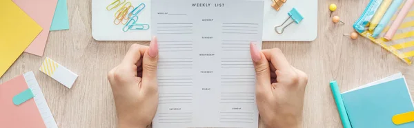 Cropped view of woman holding weekly list over wooden table with laptop and stationery — Stock Photo