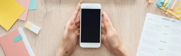 Vista recortada de la mujer sosteniendo el teléfono inteligente sobre la mesa de madera con el ordenador portátil, lista semanal y papelería - foto de stock