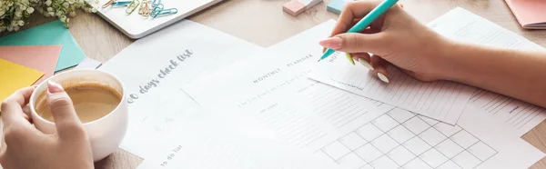 Vista ritagliata della donna che tiene penna e tazza di caffè in mano, annotazioni di scrittura nei pianificatori, seduto dietro il tavolo di legno con fiori e articoli di cancelleria — Foto stock