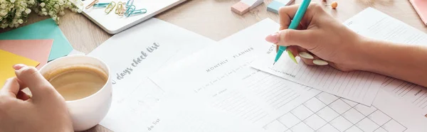 Vue recadrée de la femme tenant stylo et tasse à café dans les mains, écrivant des notes dans les planificateurs, assis derrière une table en bois avec des fleurs et de la papeterie — Photo de stock