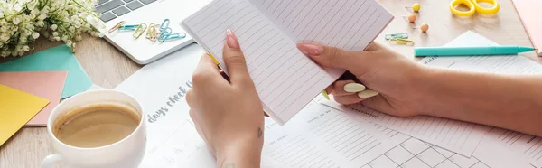 Vista ritagliata della donna che tiene il blocco note in mano, seduta dietro il tavolo di legno con tazza di caffè bianco e cancelleria — Foto stock